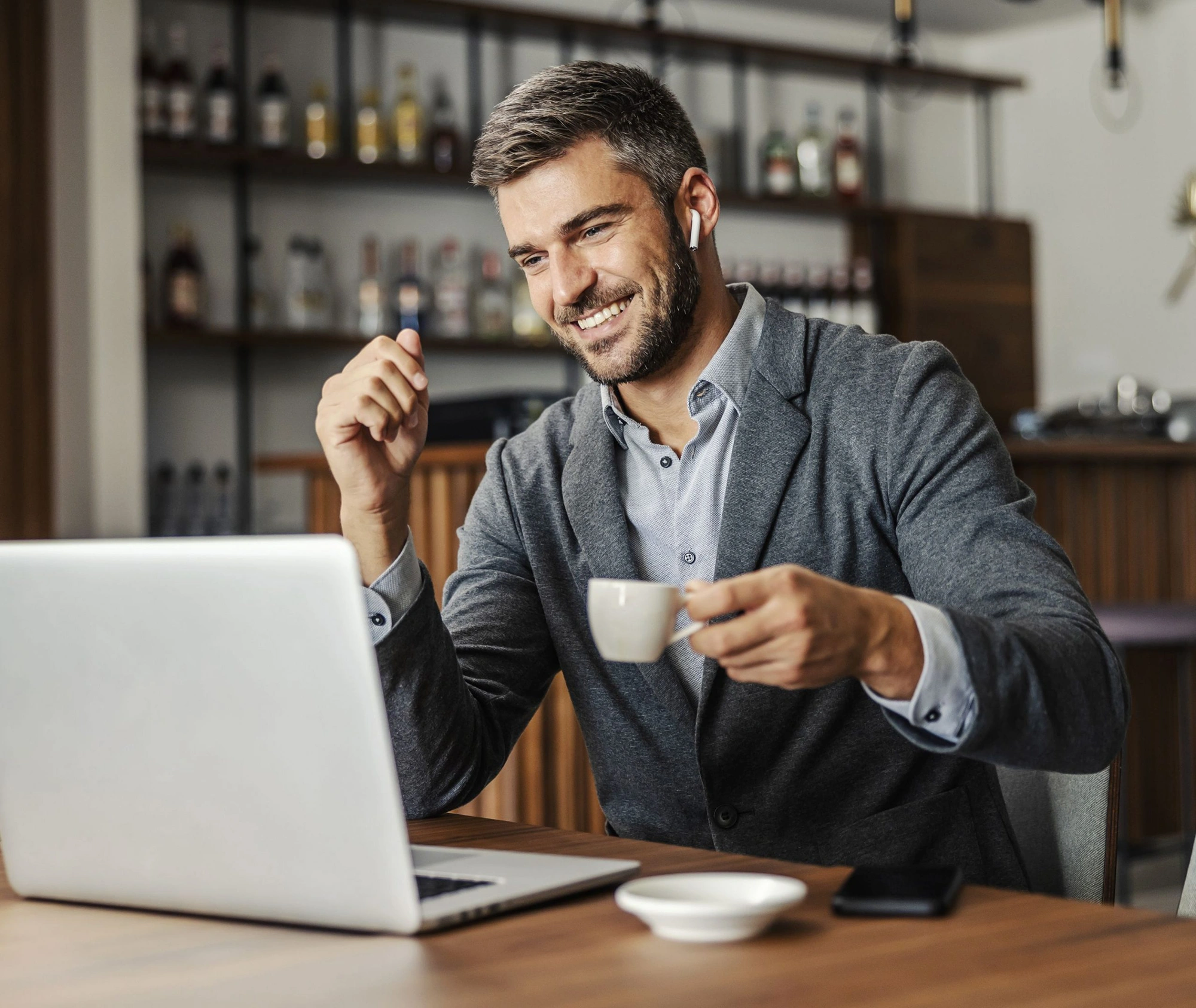 Uomo sorridente in videocall davanti al laptop in un bar moderno, tiene in mano una tazzina di caffè e indossa auricolari wireless.