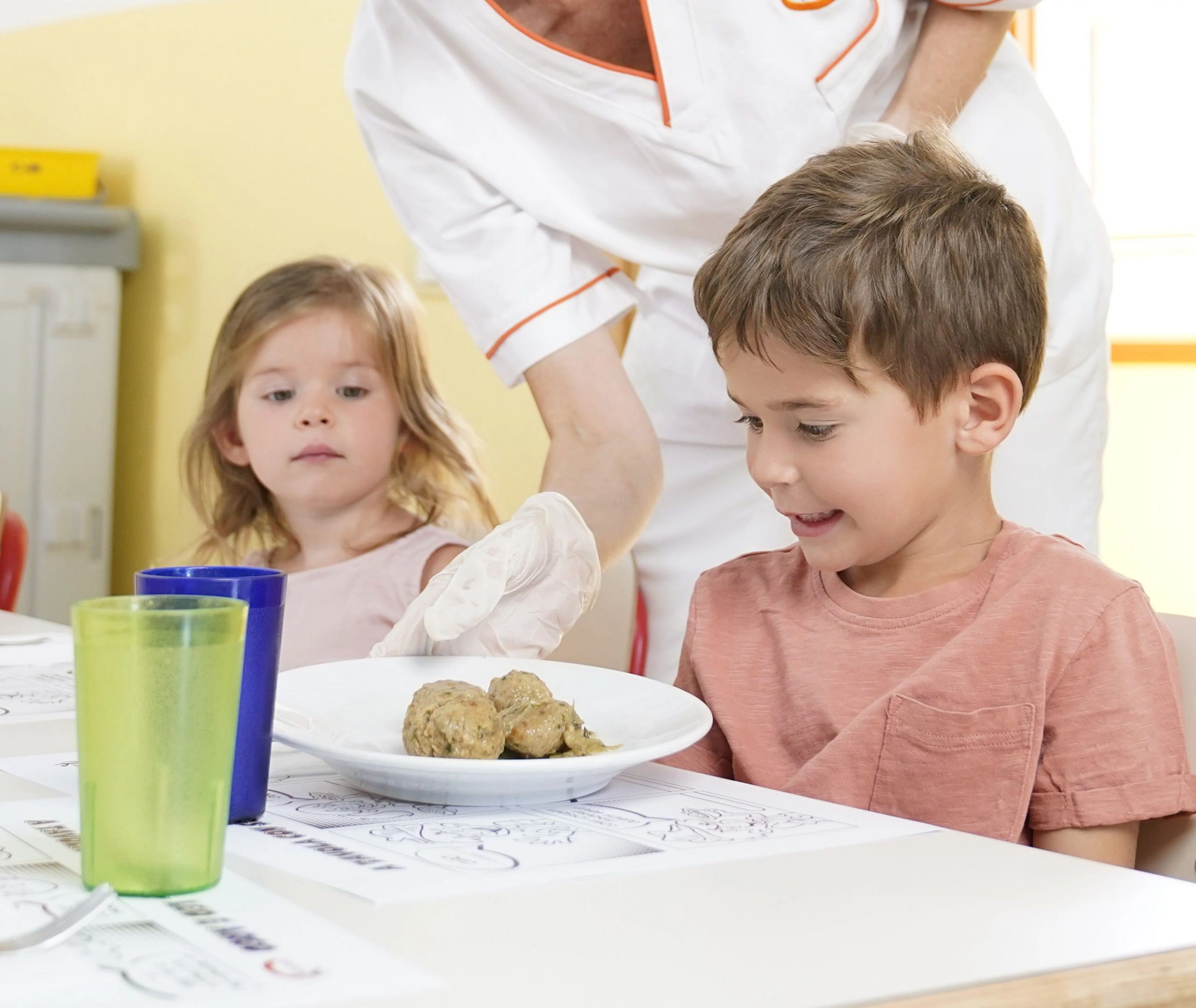 Operatrice scolastica serve un piatto con polpette a un bambino sorridente seduto a tavola insieme a una bambina, durante il pranzo in mensa scolastica.