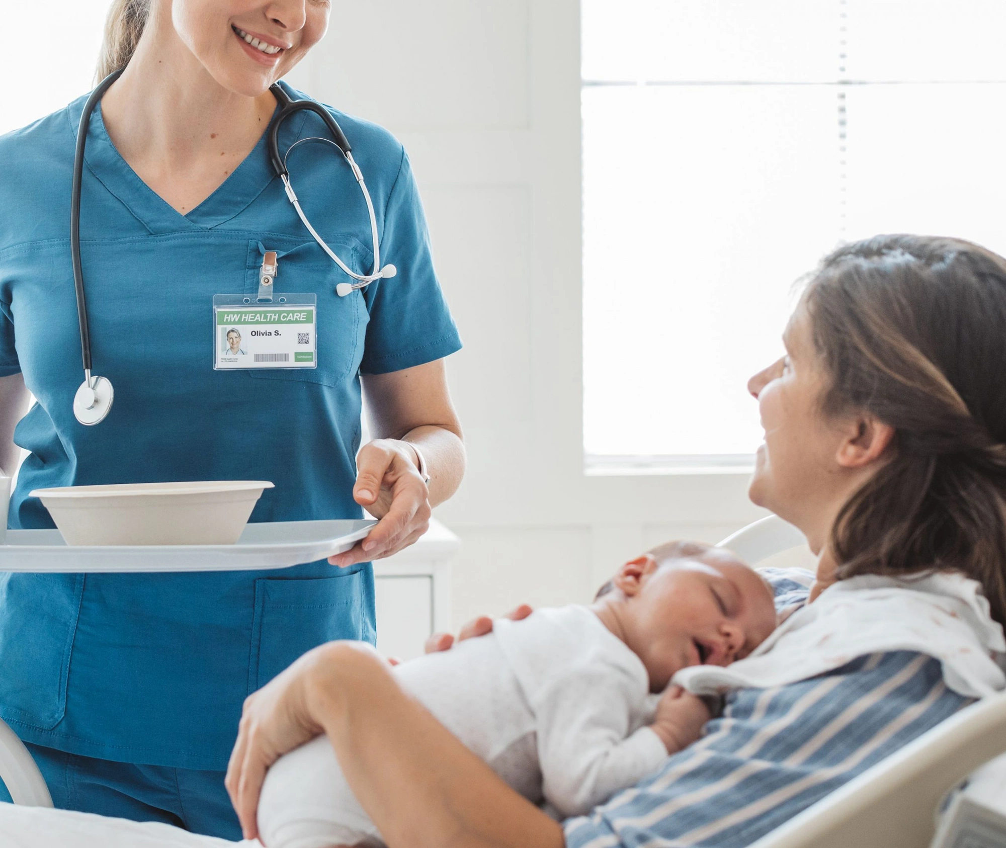 Infermiera sorridente in divisa blu consegna un vassoio con il pasto a una madre che tiene in braccio il suo neonato addormentato, in una stanza luminosa di un reparto ospedaliero.